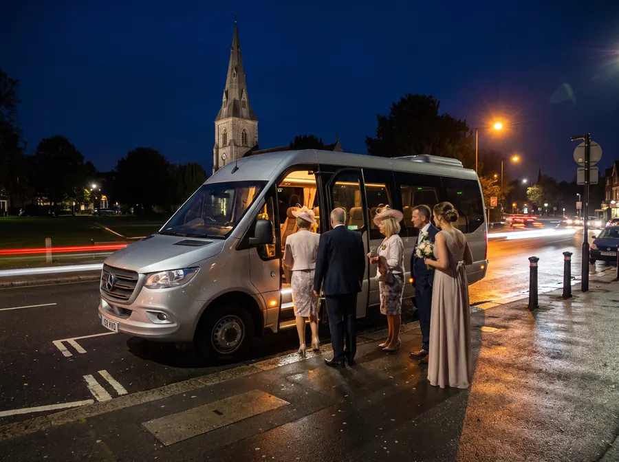A silver minibus at night on the road beside an Ealing parish church, a wedding party in hats and formal dress boarding, wet tarmac reflecting orange streetlights